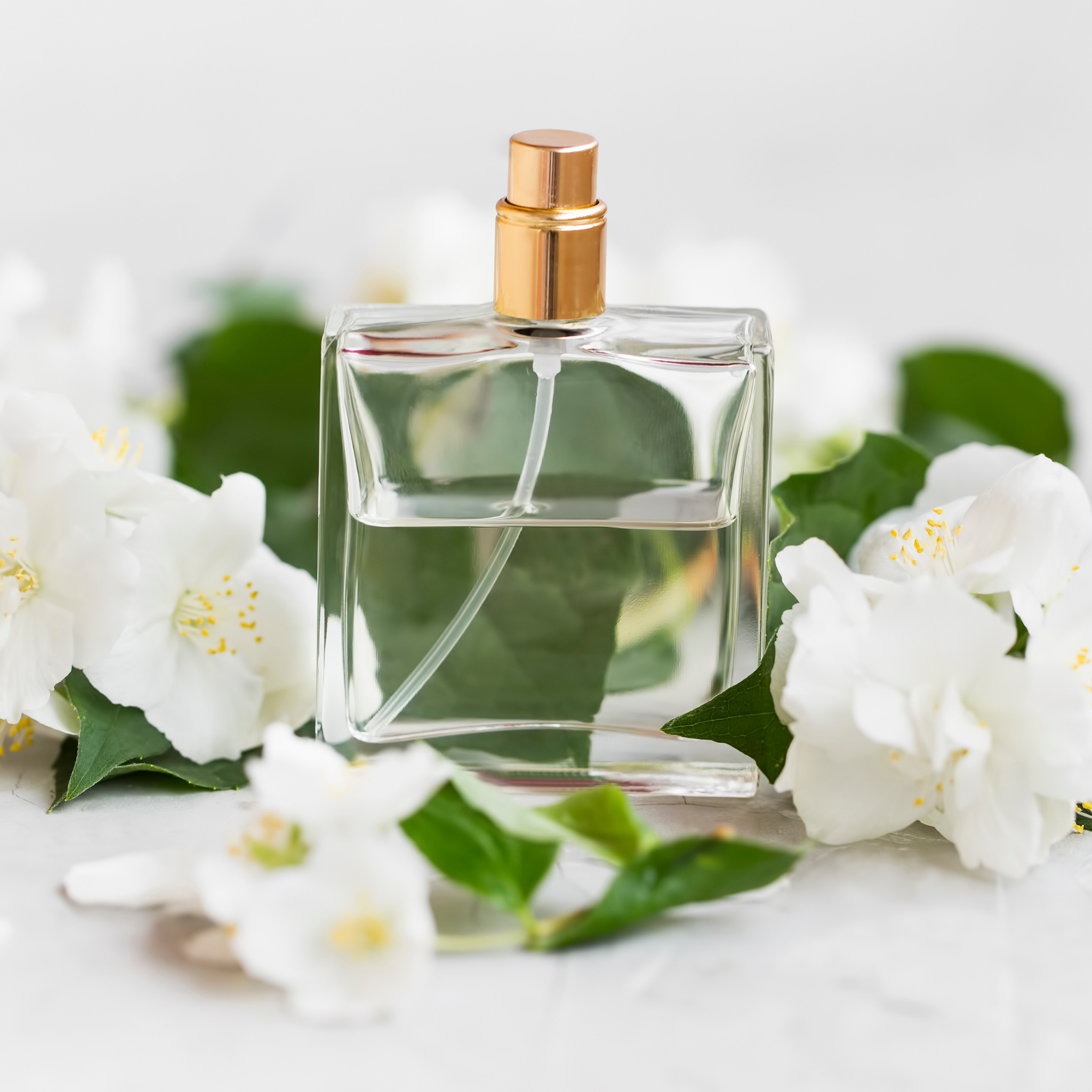 Clear perfume bottle with gold cap surrounded by white flowers and green leaves on a light background